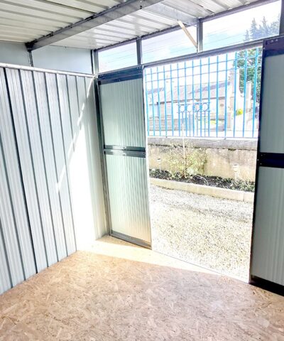 A view looking out towards the door of the pent shed. The window above the door and the open doors are allowing the strong sunlight to piur in and cast a shadow on the side wall