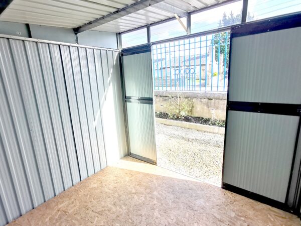 A view looking out towards the door of the pent shed. The window above the door and the open doors are allowing the strong sunlight to piur in and cast a shadow on the side wall