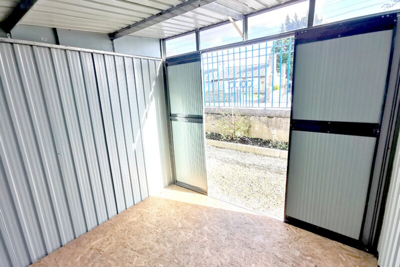 A view looking out towards the door of the pent shed. The window above the door and the open doors are allowing the strong sunlight to piur in and cast a shadow on the side wall