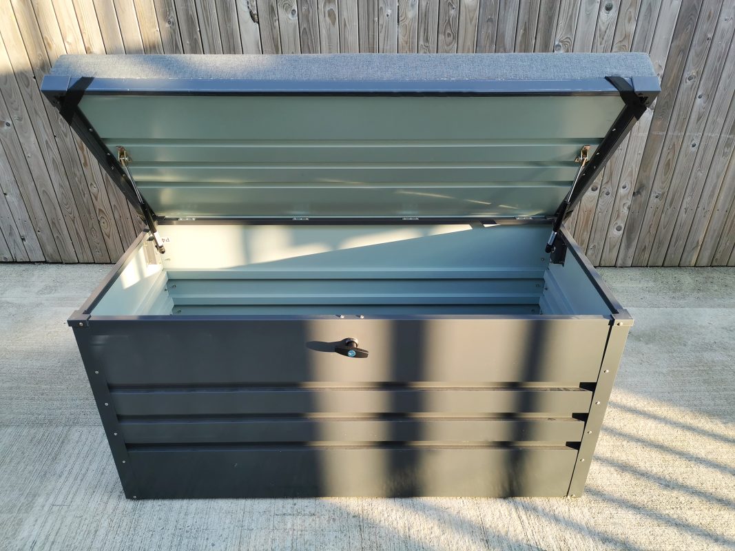 The Garden Storage Box from Sheds Direct Ireland. The box has it's hinge open and you can see inside. The unit is the 400L version and it has a grey cushion on top of the open hinge. The cushion is thick. The wall behind is entirely wooden and the sun is rising on the scene, casting long shadows.