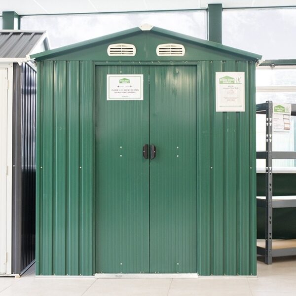 The 6ft x 5ft metal shed in the Sheds Direct Ireland Showroom. There are grey, tall shelves to the right and another shed just out of frame to the left. The 6ft x 5ft Steel Shed is green, with a sign on it that reads 'these doors slide, do not pull'
