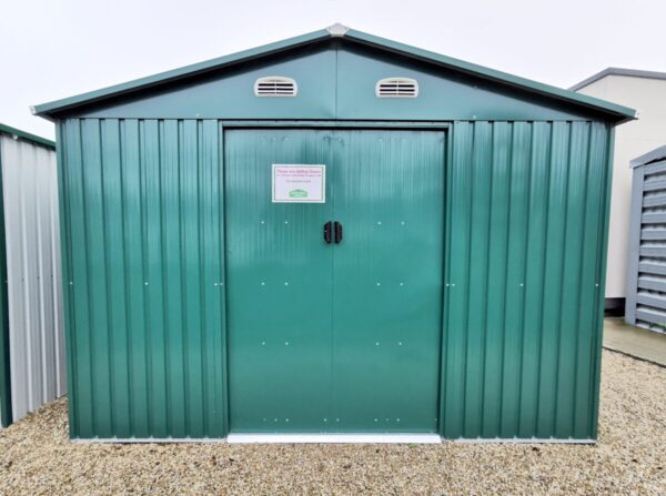 The 10ft x 10ft Shed in green on the grounds of the Sheds Direct iRELAND SHOWRROM IN NORTH DUBLIN. The base is a golden brown and the sky is bright blue. The shed is sturdy looking, the doors are closed and there is a pricelist stuck to the door.