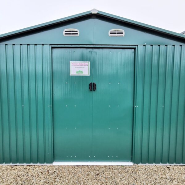 The 10ft x 10ft Shed in green on the grounds of the Sheds Direct iRELAND SHOWRROM IN NORTH DUBLIN. The base is a golden brown and the sky is bright blue. The shed is sturdy looking, the doors are closed and there is a pricelist stuck to the door.
