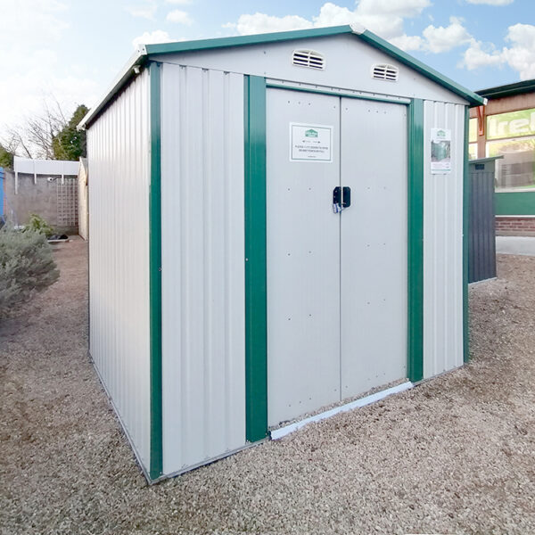 The 8ft x 6ft steel garden shed on the Sheds Direct Ireland lot. It's a white-white colour with green trim. It stands on cobblestones that are goldish in colour and the showroom walls are visible behind it.