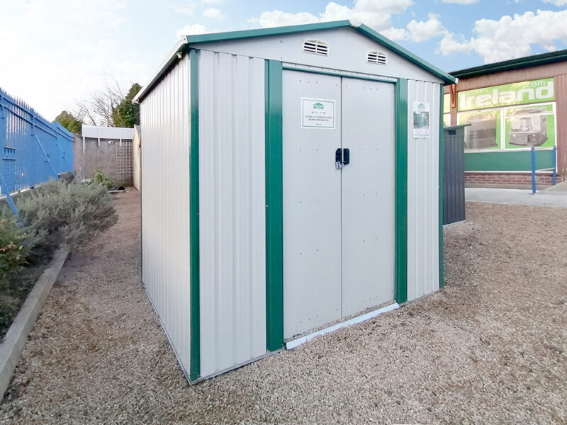 The 8ft x 6ft steel garden shed on the Sheds Direct Ireland lot. It's a white-white colour with green trim. It stands on cobblestones that are goldish in colour and the showroom walls are visible behind it.