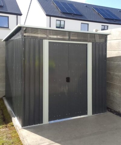 A photo of the pent shed in a customers garden. It sits on a pale grey slab of concrete with green grass to the left of the frame. There is a towel on the washing line above the grass also