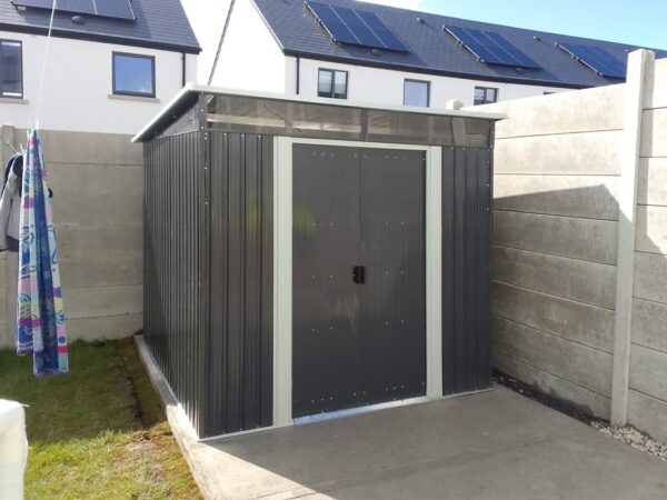A photo of the pent shed in a customers garden. It sits on a pale grey slab of concrete with green grass to the left of the frame. There is a towel on the washing line above the grass also