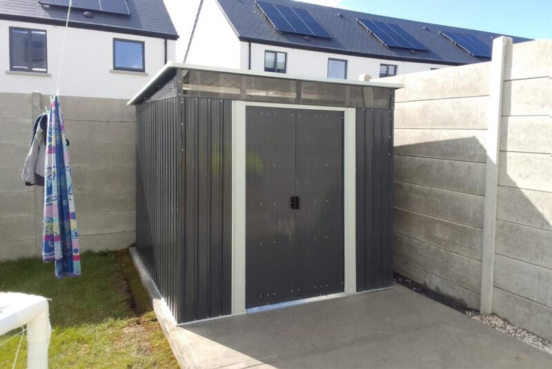 A photo of the pent shed in a customers garden. It sits on a pale grey slab of concrete with green grass to the left of the frame. There is a towel on the washing line above the grass also