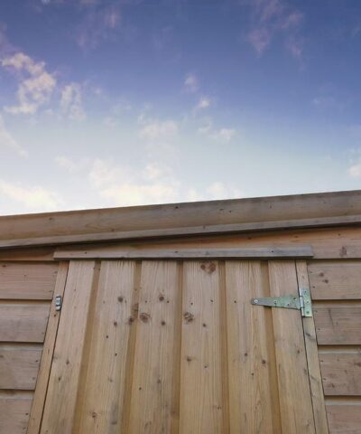A close up of a wooden cabin shed. The pent roof is sloping down from the right to the left. The top of the wooden door is visible. The sunrise is occurring behind the shed - it is fading from bright blue to dark blue as the sun rises. There is nothing else in the image.