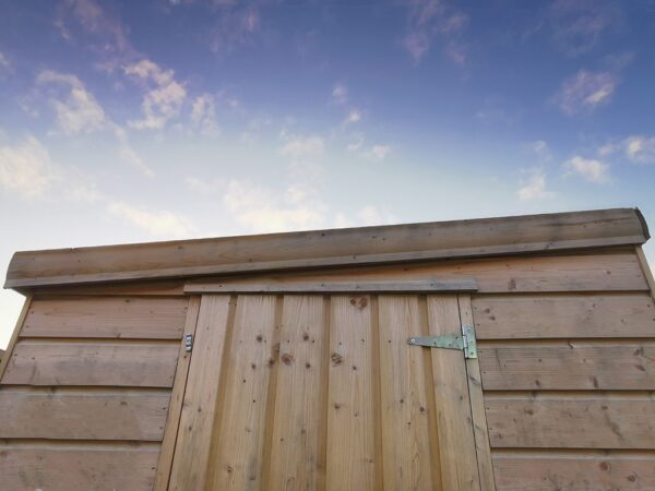 A close up of a wooden cabin shed. The pent roof is sloping down from the right to the left. The top of the wooden door is visible. The sunrise is occurring behind the shed - it is fading from bright blue to dark blue as the sun rises. There is nothing else in the image.
