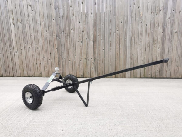 A black trailer dolly with silver, circular ball attachment. The trailer dolly is in a flat position with the handle pointing upwards at a 30 degree angle. It's sitting on a pale paving slab with a wooden wall behind it.