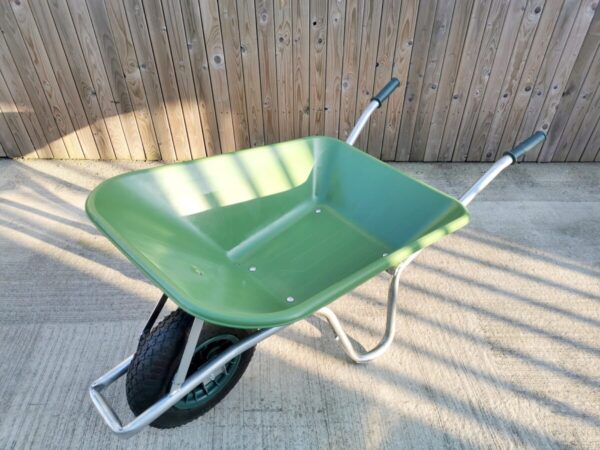 The wheelbarrow as seen from above. It is green with steel handles, a square wheel protector and black handles and tyre