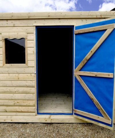 The wooden cabin shed with the door open as seen from the front. The internal wooden floor is visible, but one window is hidden behind the open door. The inside of the door has a blue lining which is for ventilation and temperature control. The shed is on hard gravel and placed beside other sheds. The sky is blue above.