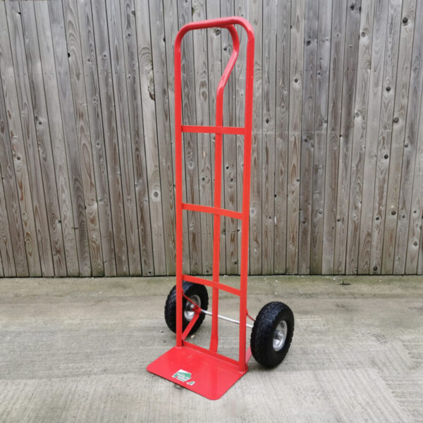 A Red sack truck from Sheds Direct Ireland against a wooden wall. The trolley is large, about 5 foot tall. It has two large outer bars which run from the base to the curved handle at the top. There is an internal bar, which is connected to the external ones by three horizontal bars. They are all red. There is a P shaped handle at the back which curves outward. The wheels at the bottom are thick and black.