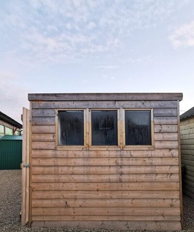 The side of the cabin shed in the showroom. It is a wide angled shot and the sky has some small clouds in the distance. The clouds are orange due to the sunrise. The shed has three black windows which have some morning dew on them. The wood is a pale colour and the wooden slats are horizontal