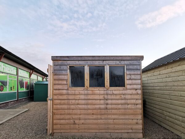 The side of the cabin shed in the showroom. It is a wide angled shot and the sky has some small clouds in the distance. The clouds are orange due to the sunrise. The shed has three black windows which have some morning dew on them. The wood is a pale colour and the wooden slats are horizontal