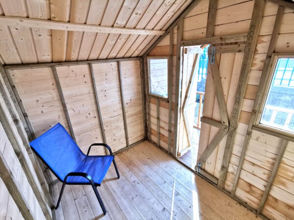 A view of the inside the wooden chalet shed. The shed is a pale brown colour with darker support beams. There is a blue chair in the middle of the shed.