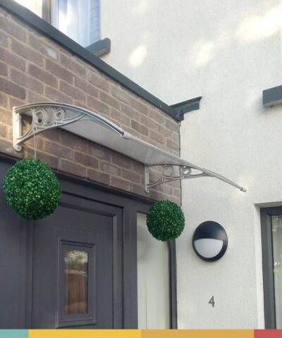 A customers photos showing a door canopy installed above a greay door onto some redbrick. There are two circular flower beds suspended from the door canopy and above the canopy there is a clear blue sky.