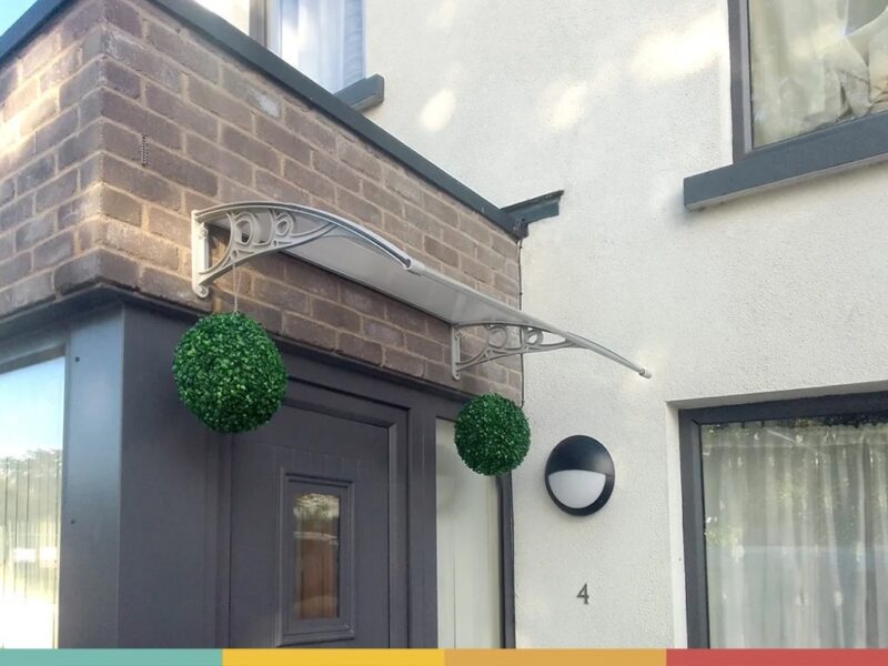 A customers photos showing a door canopy installed above a greay door onto some redbrick. There are two circular flower beds suspended from the door canopy and above the canopy there is a clear blue sky.