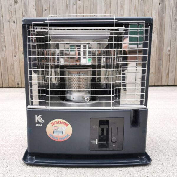 The Tosai / Kero360 Heater against a wooden wall and sitting on a concrete base. It's a large boxy unit, with a black bottom haldf and a metallic upper half. In the metallic half, a silver, tubular burner can be seen. A large yellow sticker below this is emblazoned with a sign that says '3000W'
