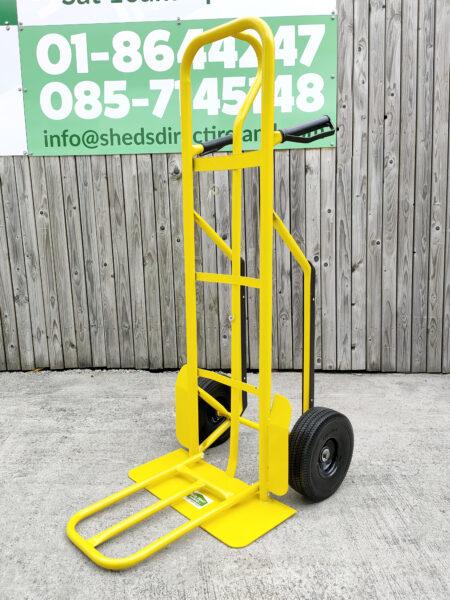 The side view of the bright yellow industrial hand truck. It's against a wooden wall and the extendable base plate has been folded out