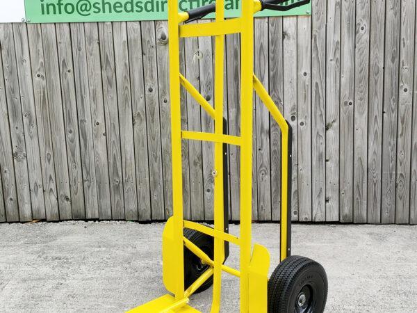 The side view of the bright yellow industrial hand truck. It's against a wooden wall and the extendable base plate has been folded out