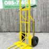 The side view of the bright yellow industrial hand truck. It's against a wooden wall and the extendable base plate has been folded out