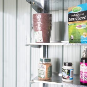 Metal Took Shelves mounted on the inside of a steel shed. There is a blue and green box of bird seed, a pack of plastic plant pots, an orange box of grass seed, a small white jar of 'loco coco' and a purple, wine-bottle shaped bottle of 'Orchid Mister' on the shelves. The ehslves themselves are a patchy steel.