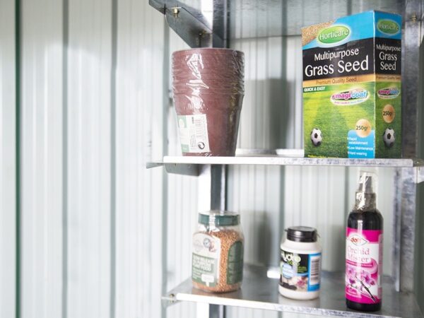 Metal Took Shelves mounted on the inside of a steel shed. There is a blue and green box of bird seed, a pack of plastic plant pots, an orange box of grass seed, a small white jar of 'loco coco' and a purple, wine-bottle shaped bottle of 'Orchid Mister' on the shelves. The ehslves themselves are a patchy steel.