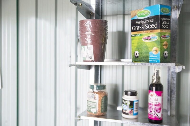 Metal Took Shelves mounted on the inside of a steel shed. There is a blue and green box of bird seed, a pack of plastic plant pots, an orange box of grass seed, a small white jar of 'loco coco' and a purple, wine-bottle shaped bottle of 'Orchid Mister' on the shelves. The ehslves themselves are a patchy steel.