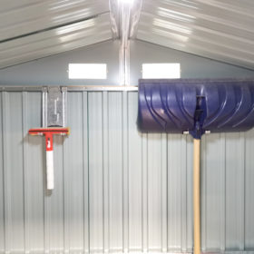 A wide angle picture of the inside of a shed with the focus on two took hooks on the back wall. One is supporting a large, blue snow shovel. The other is supporting a small, red squeegee. The hooks are 30cm long, 10cm wide and they have a picture of a shovel indented into the,