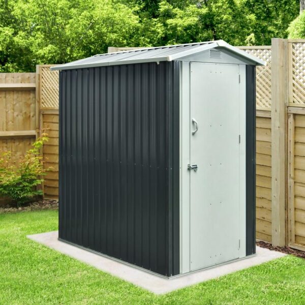 The New 4x6 Steel Garden Shed from Sheds Direct Ireland. It's a dark grey in colour overall, with a lighter grey door and peaked roof. There is a handle on the door as well as a bolt-lock. There is a wooden wall behind the unit and to it's right hand side. It sits on a solid concrete base and there's a large tree behind it too!