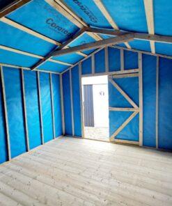 The internal view of a wooden shed, looking outwards. The light is pouring in the door and the walls have . abright blue membrane lining on them. There are thick wooden slats and two windows on the right hand side of the shed as we view it.