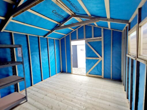The internal view of a wooden shed, looking outwards. The light is pouring in the door and the walls have . abright blue membrane lining on them. There are thick wooden slats and two windows on the right hand side of the shed as we view it.