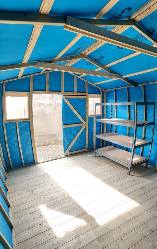 A vertical image taken inside the taller wooden shed. The sides are warped, as a fish-eye lens has been used. The walls inside the shed are blue, the floor is a pale wood colour and light is pouring in the door. Outside the door, only a grey wall is visible