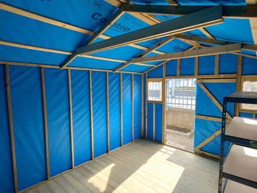 inside the taller wooden shed. The walls are a bright blue, as they are lined with membrane material and the floor is a pale wooden colour. The roof is very high up and it's reinforced with thick, darker wooden bars.