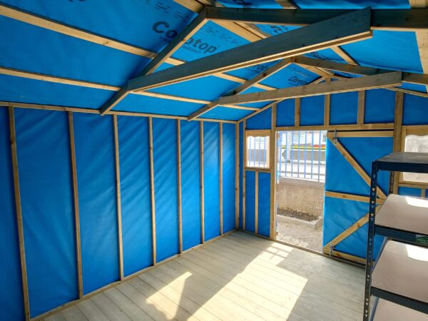 inside the taller wooden shed. The walls are a bright blue, as they are lined with membrane material and the floor is a pale wooden colour. The roof is very high up and it's reinforced with thick, darker wooden bars.