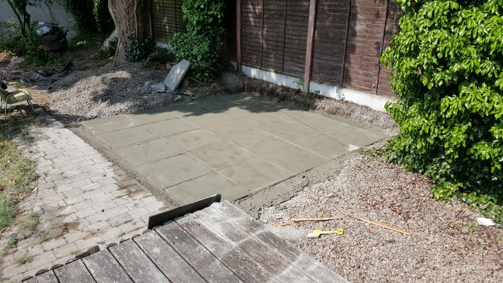 A Solid level base made of slabbed concrete against a garden wall. There is a bush nearby but which is not encroaching on the base