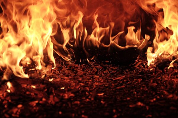 A close up of a burning fire, which is dark in the foreground with lapping yellow flames on top. Above, the yellow flames are becoming orange and brown and there is a large amount of smoke. Photo by Raquel Raclette