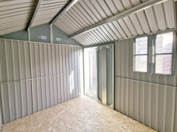 Inside the 10ft x 7.5ft steel cottage shed. The view is from the end opposite door. The doors are open the window is letting light flow in and the apex is visible too. The support frame beams are shiny metal and the walls are a matte-effect metal. The floor is a plywood.