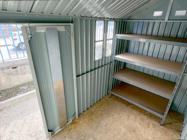 Inside the 10ft x 6ft Steel Cottage Shed from Sheds Direct Ireland. The walls are a pal grey colour, there is a wooden ply floor and a grey and brown large shelving unit inside.