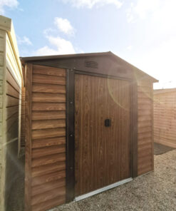 Woodgrain Metal Sheds in our Finglas Showroom. They are brown and have a faux-woodline appearance. They are standing on a concrete ground and the sun is setting behind them to the righ,creating a golden sky and a camera-lens flare onto one of the sheds