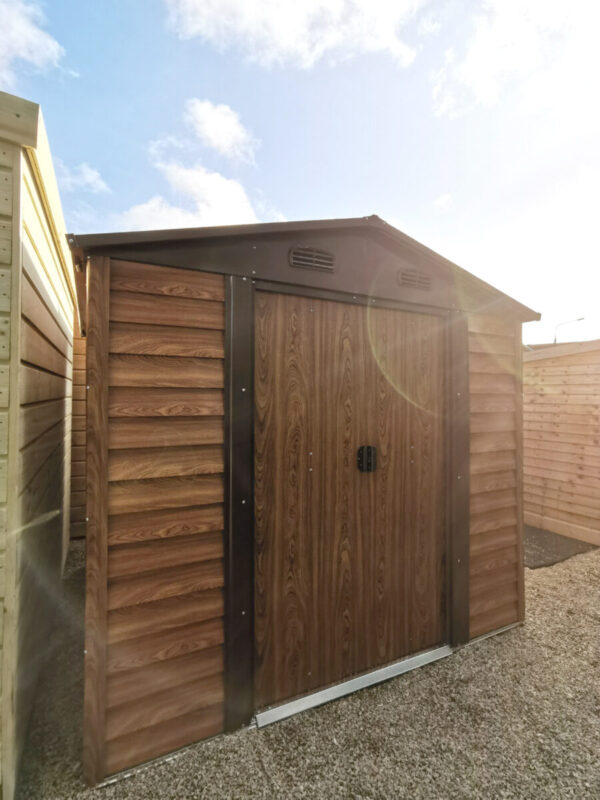 Woodgrain Metal Sheds in our Finglas Showroom. They are brown and have a faux-woodline appearance. They are standing on a concrete ground and the sun is setting behind them to the righ,creating a golden sky and a camera-lens flare onto one of the sheds