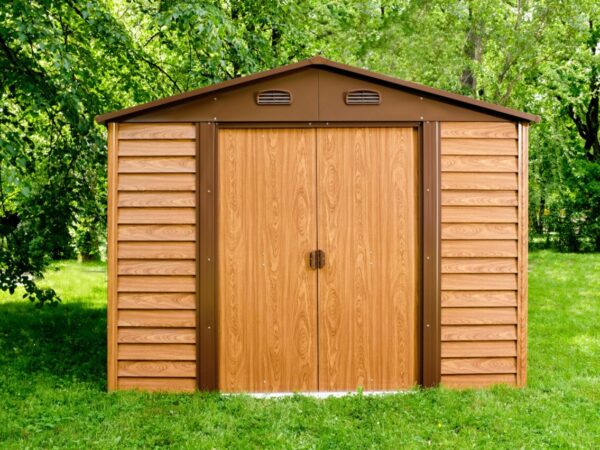 The 9ft x 8ft woodgrain metal shed under a willow tree in a large garden. The shed is oak brown, with chocolate brown flashings around the door and vents. The shed is tall and wide and it casts a long shadow.