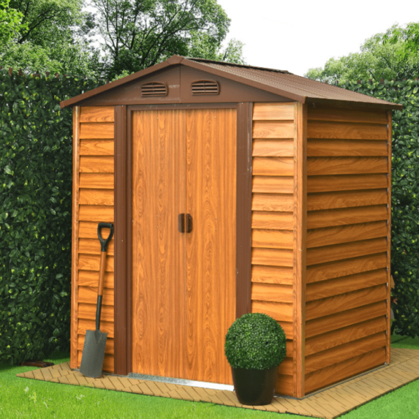 The 8ft x 6ft Wood Grain Steel Shed is sitting in a garden, close to a hedge. It is on a yellow-stone concrete base with hedges to two sides of it. There is a plant pot with a circular shrubbery in it and a shovel to the front of the shed
