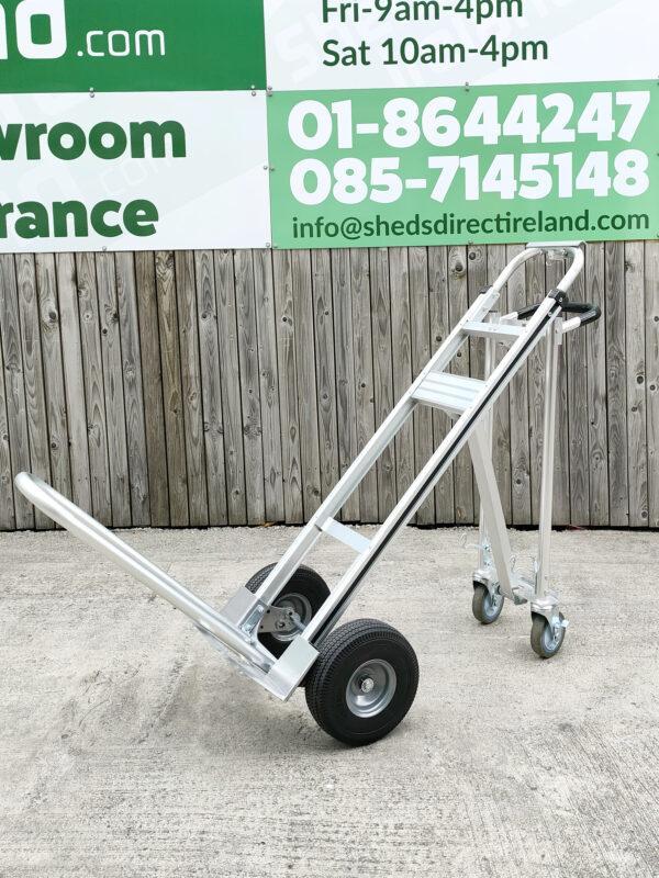 the three in one hand truck in the horizontal position. The extendable arm is also fully extended. The unit is a shiny grey metal and it stands against a wooden wall
