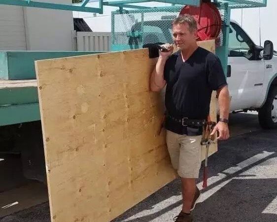 A man carrying a large plywood board using the panel carrier. He is holding it at shoulder height and has unloaded it off the side of a white truck beside him.