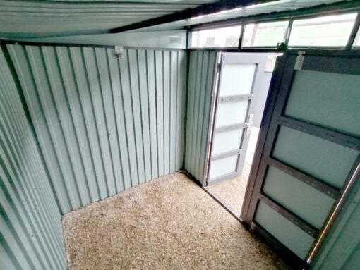 Inside the Premium Pitched Steel Garden Shed. It has no floor and the pebblestones are visible as the base. The door is open and light pours in through it and also from the large window above the door