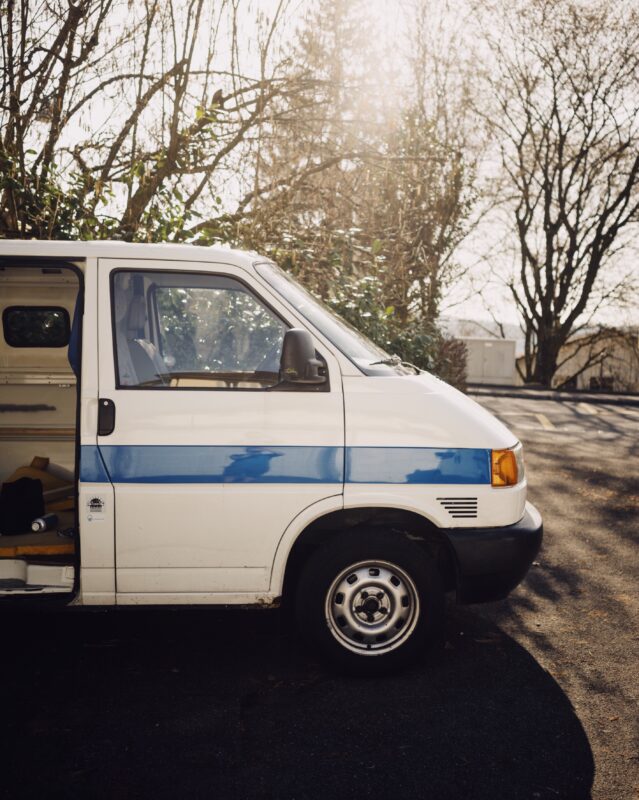 A white transport can with the door open is backlight by a warm wintery sky. The trees behind are bare and the van is empty of people. The van is white with a blue strip along the side. 