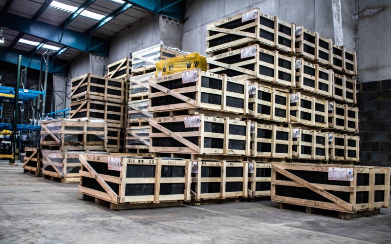 An inside view of a large warehouse. There are hundreds of pale brown pallets stacked high with thickly wrapped, black boxes. There light above is fluorescent and it casts long shadows on the stuff below.